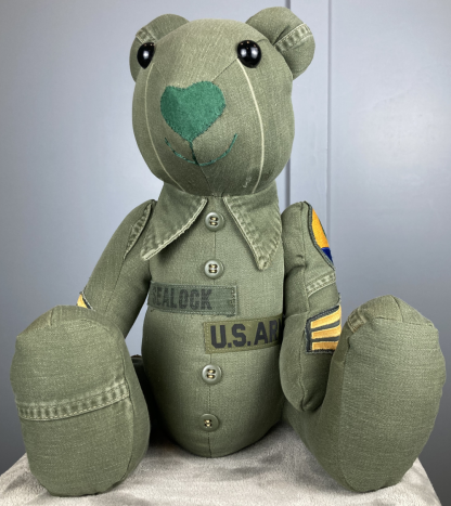 bear with heart shaped nose, color army green from an army jacket. patches on the arms of the jacket and name patches on the stomach. buttons going down the center and the color to the jacket on the neck of the bear.